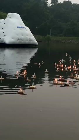 The video shows a serene body of water, likely a lake or pond, with a large white inflatable structure on the left side of the frame. Floating on the water's surface are numerous small rafts, each holding a lit candle. These floating candles create a soft, flickering glow, reflecting on the dark water. The background features a lush green forest covering a hillside, and the sky appears overcast, suggesting it may be early morning or late evening. There are no visible people or animals in the immediate foreground, lending a peaceful and contemplative atmosphere to the scene.