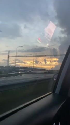 The video shows a view from inside a moving car, looking out the passenger window at dusk. The sky is filled with dramatic clouds, with a warm orange and yellow sunset peeking through. There is a highway visible in the foreground with some streetlights and other vehicles. A reflection of what appears to be fire or a bright light source can be seen on the car window. The overall impression is a scenic drive during sunset.