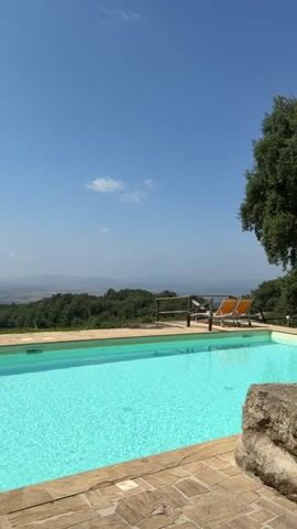 The video shows a swimming pool with bright turquoise water, set against a backdrop of rolling green hills and a hazy blue sky. Two orange lounge chairs are visible by the edge of the pool, and a large, rough-hewn rock is in the foreground. The pool deck is made of stone tiles. A tree is visible on the right side of the frame. The overall impression is one of a serene, outdoor setting on a clear day.