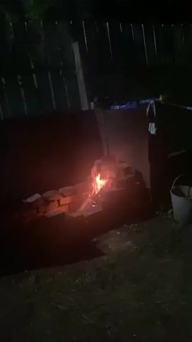 A small fire burns in a makeshift brick fireplace at night. The fire is contained within a U-shaped structure built from red bricks. Flames flicker and rise from the center of the fireplace, casting a warm glow on the surrounding area. The background is dark, with a fence visible in the upper left portion of the frame. A white bucket sits to the right of the fireplace. The ground is covered in grass and dirt.