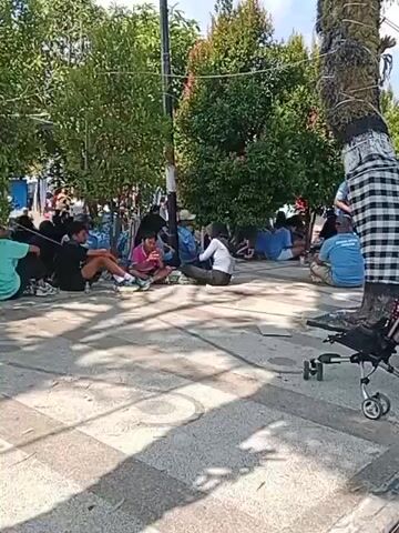A group of people are sitting on the ground in a shaded area, possibly a park or a public space. There are trees and foliage in the background, suggesting an outdoor setting. Some individuals are dressed casually, while others are wearing blue shirts, possibly indicating they are part of a group or organization. One person is seen using a mobile phone, and another appears to be eating. A stroller is visible in the foreground, along with a tree wrapped in a checkered cloth. The ground is paved with cobblestones and has some patterned markings. The lighting suggests it is daytime, with sunlight filtering through the trees. The overall impression is one of a gathering or waiting period.