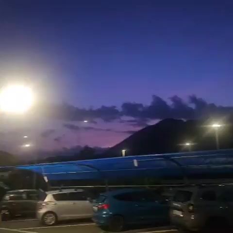 The video shows a parking lot at dusk with a bright light source resembling the moon or a streetlamp in the upper left quadrant. Several cars are parked, including a silver hatchback and a blue hatchback. A person is standing near the open trunk of a gray sedan. The sky is a deep purple with some clouds. In the distance, hills or mountains are visible. The scene appears to be outdoors, possibly in a semi-rural or suburban area. A blue tarp structure is present over some of the parking spaces. A crescent moon is visible in the sky in the latter part of the video.