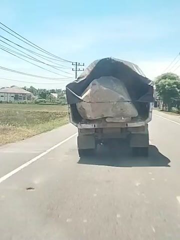 A truck is driving down a paved road.  The truck's bed is filled with a large rock that is partially covered by a dark tarp. Exhaust fumes billow from the truck as it moves.  The environment includes houses, grass, trees, and telephone poles alongside the road under a bright blue sky. A person on a motorcycle passes the truck.