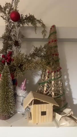 The video shows a close-up view of Christmas decorations displayed on a shelf. The decorations include various figurines such as Christmas trees, gnomes, angels, a snowman, and a Santa Claus. There are also natural elements like pinecones, berries, and branches, along with festive ornaments like red balls and glittery accents. A small, handmade wooden house is also visible. The lighting is soft and warm, suggesting an indoor setting. The camera pans slowly across the shelf, showcasing the different decorative items.