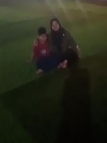 A blurry video shows a woman in a hijab and a child sitting on a green surface, likely grass or artificial turf, in dim lighting. The woman has her arm around the child, and they appear to be looking at the camera. The child is wearing a red shirt and blue pants, while the woman is wearing a dark top and pants. A shadow of a person is visible on the ground to the left.