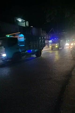 The video shows a nighttime street scene with several vehicles. In the foreground, a dark-colored truck with blue lights beneath its chassis is moving away from the camera. To the right, a lighter-colored truck with flashing blue lights on its roof and yellow headlights is moving towards the camera. The road appears to be wet, reflecting the vehicle lights. Buildings are visible on the left side of the street, and trees are present on the right. A motorcycle is parked on the right side of the road. The overall impression is of a street with active traffic at night.