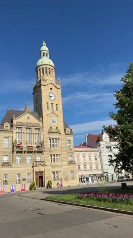 The video shows a stone building with a prominent clock tower, set against a bright blue sky with scattered clouds. The building appears to be a town hall or similar civic structure, with architectural details including arched windows, balconies, and decorative carvings. Two large clocks are visible on the tower. In the foreground, a paved area transitions to a strip of grass with flowering plants. A few people are walking in the background, and trees are visible on the right side of the frame. The overall impression is of a sunny day in a European town square.