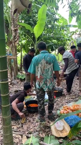 A group of people are gathered in a wooded area, possibly a jungle or forest, with lush green foliage and tall trees. The ground is covered in dirt and fallen leaves. Several individuals are standing and observing while others are engaged in activities. In the foreground, one person is kneeling and appears to be preparing food in a large pot. Another person stands nearby, holding a bottle. In the midground, a spread of food, including what appears to be cooked shrimp or prawns, is laid out on banana leaves. Several children are present, some standing and interacting. In the background, more people are visible, and a motorcycle is parked. The overall scene suggests an outdoor gathering or meal preparation in a natural setting.