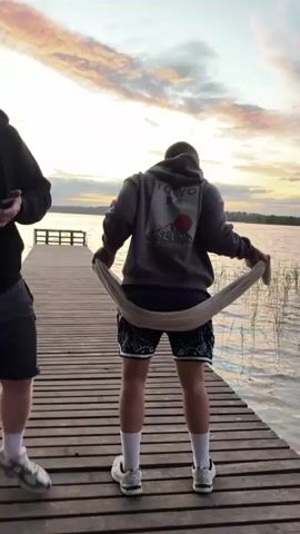 Two men stand on a wooden dock by a lake at sunset. The man on the left wears a black zip-up hoodie and gray shorts. He is recording video on his phone and smiling. The man on the right wears a gray hoodie, patterned shorts, and white shoes. He has a towel around his waist. In the background, the sky is a mix of orange, pink, and gray hues. The lake is calm with a few reeds visible near the dock.