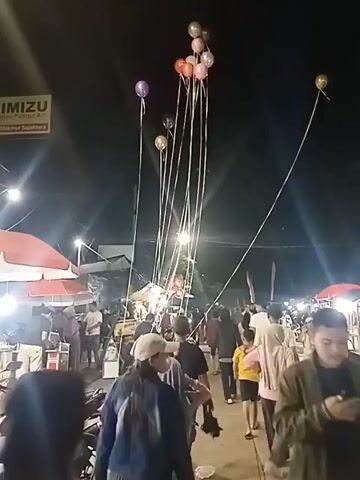 The video shows a nighttime scene at what appears to be an outdoor market or festival. A group of people are walking down a path, and in the foreground, a person wearing a red jacket is walking away from the camera. Overhead, numerous balloons are tethered by long strings and rise into the dark sky. Some of the balloons are clustered together, while others are more spread out. In the background, there are stalls with vendors, umbrellas, and lights, indicating commercial activity. The overall atmosphere seems lively and social.
