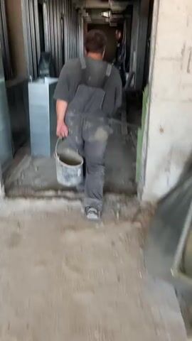 A construction worker in gray overalls is walking down a hallway, carrying a bucket in their right hand. The hallway is under construction, with debris and materials scattered on the floor. Other construction workers can be seen in the background. The lighting is dim and appears to be artificial. The camera is following the worker from behind and to the side.