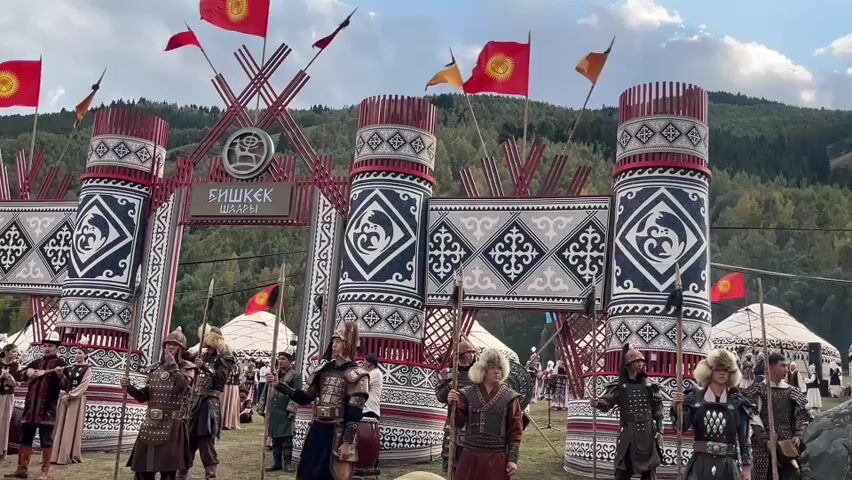 The video shows an outdoor scene with a large, ornate gate at the center, decorated with traditional patterns and flags. A group of people dressed in historical armor and fur hats stand in front of the gate, holding spears. Some of these individuals appear to be actors or performers. In the background, several yurt-like structures are visible, suggesting a nomadic or cultural gathering. A statue of a snow leopard sits on a rocky outcrop to the right of the gate. The environment appears to be a grassy field with a forested hillside in the distance, under a partly cloudy sky. Several people in the foreground are holding up mobile phones, likely recording the event.