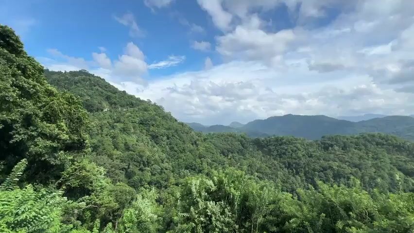 The video shows a scenic view of a mountainous landscape covered in lush green forests under a partly cloudy sky. The mountains recede into the distance, their peaks appearing less defined due to the atmospheric perspective.  The foreground features dense vegetation, suggesting the vantage point is at a high elevation.  The sky is a mix of blue patches and white, puffy clouds.  The video appears to be taken during daylight hours. A vehicle can be heard in the distance.