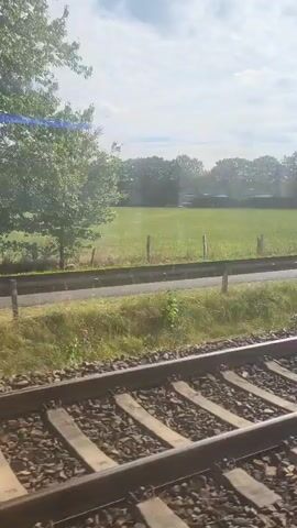 A short video shot from a train window. The train tracks are in the foreground. In the middle ground, there is a fence, a road, and a grassy field with a cell tower visible in the distance. The background consists of trees and a partly cloudy sky. The train appears to be moving.