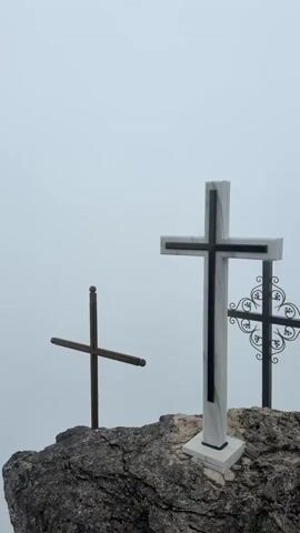 The video shows three crosses on a rocky outcrop with a foggy or overcast sky in the background. The largest cross is in the foreground and is made of white marble with black accents. To the left is a simpler wooden cross with a thin pole. To the right, slightly behind the large cross, is a dark metal cross with decorative scrollwork. The rock appears rough and natural. There is no visible activity or interaction, and the focus is on the static arrangement of the crosses in the environment.