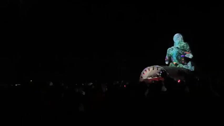 The video opens with a wide shot of a nighttime scene, dominated by darkness. In the background, a large, illuminated sculpture of a human-like figure is visible. The sculpture is lit with vibrant red and purple lights, giving it an otherworldly appearance. It sits atop a stage or platform structure, which also has lights. In front of the stage, a crowd of people is gathered, their forms mostly silhouetted against the light. Some individuals in the crowd appear to be holding up their phones, likely recording or taking pictures. Two red lights are visible in the sky above the stage, possibly pyrotechnics or stage lighting. The overall impression is of an outdoor concert or festival event.