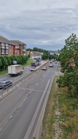 The media file shows a view of a road in Canterbury, United Kingdom. The scene is captured from an elevated position, likely a bridge or overpass, looking down onto a two-lane road with traffic.

The primary subjects are vehicles on the road. These include cars of various colors, a delivery truck with a white trailer, and a larger truck with a blue and white cab. The road itself is asphalt, marked with white lines indicating lanes and a central divider. A metal railing runs along the side of the road, separating it from a grassy verge with wildflowers and trees.

On the left side of the road, there are brick buildings with multiple stories, likely apartment blocks or offices, surrounded by trees. On the right side, there are houses with pitched roofs and more trees, including a large leafy tree in the foreground. In the distance, more buildings and trees are visible under a cloudy sky.

The activity taking place is traffic moving along the road. Vehicles are seen in motion in both directions. The time of day appears to be daytime, given the overall illumination, but the cloudy sky suggests it might be overcast. There are no people clearly visible in the media file.

Notable details include the overcast weather conditions, indicated by the gray and cloudy sky. The location is identified as Canterbury, United Kingdom. No text is clearly legible in the provided frame. The overall mood is neutral, documenting a typical street scene.