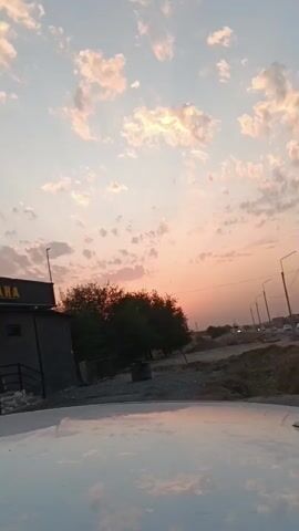 This is a vertical, handheld video showing a street scene during sunset. The sky is filled with scattered clouds tinged with pink and orange hues. In the foreground, the hood of a white car is visible, reflecting the sky. To the left, a dark building with a sign that reads "KAATKANA" is partially visible. A few trees with dark green foliage stand behind the building and in front of the road. The road itself stretches into the distance, lined with streetlights. Several cars are driving on the road. The overall environment appears somewhat arid, with dirt and gravel visible on the sides of the road. The camera pans slightly upwards throughout the video, following the sky.