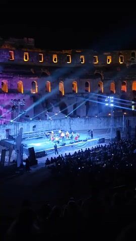 This video captures an outdoor concert held at night within the ruins of an ancient amphitheater. A stage is set up in the center, illuminated by blue and purple stage lights that cast beams across the scene. On the stage, a group of musicians, likely an orchestra, is performing. Several individuals are visible playing string instruments, such as cellos, and one person appears to be playing a keyboard or piano. A camera operator stands to the right of the stage, filming the performance. In front of the stage, a large audience is seated, facing the performers. Many audience members are holding up their phones, presumably to record or photograph the event. The background of the amphitheater shows stone walls with arched openings, illuminated from within by warm, orange light, highlighting the historic architecture. The overall atmosphere is one of a cultural event blending historical surroundings with contemporary entertainment.