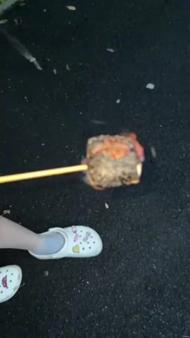 A person is holding a skewer with a marshmallow over a fire. The marshmallow is slightly burnt and smoking. The person is wearing white Crocs with cartoon characters on them. The ground appears to be asphalt or pavement, with some fallen leaves scattered on it.