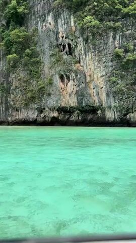 A boat with people on it passes by a large cliff face that is covered with green trees and vegetation. The water in the foreground is a bright turquoise color and appears to be very clear. The cliff is rocky and has several crevices and small caves. The boat is long and narrow and has a canopy over the back. There are several people visible on the boat, some sitting and some standing. One person is wearing a pink shirt. The overall impression is of a beautiful and serene tropical landscape.