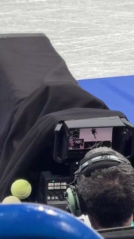A cameraman with headphones is operating a camera covered with a black cloth. The camera has a small screen showing a figure skater performing. A couple of tennis balls sit near the camera. The floor is white and blue.