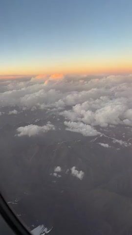 The video shows an aerial view from an airplane window. The scene is dominated by mountains partially obscured by clouds. The sky above displays a gradient of colors, transitioning from a pale blue at the top to soft oranges and pinks near the horizon, indicating either sunrise or sunset. The clouds are fluffy and white, with some areas illuminated by the warm, low-angled sunlight. The mountains below are rugged and appear to have snow-capped peaks or patches of snow. The overall impression is one of serene natural beauty viewed from a high altitude. There are no people or objects other than the natural landscape and the airplane window visible in the frame.