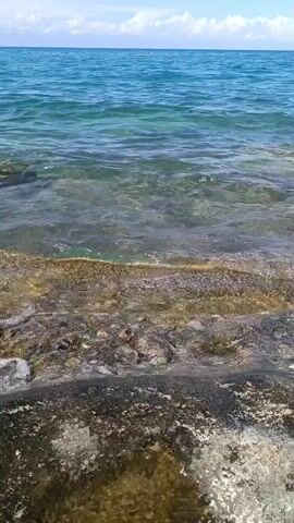 The video shows a close-up view of clear blue ocean water lapping against a rocky shore. The waves are gentle, creating ripples and foam as they reach the edge. Sunlight reflects off the water's surface, illuminating the submerged rocks and seabed. The horizon line is visible in the distance, separating the ocean from a bright sky with scattered clouds. The scene is peaceful and natural, evoking a sense of tranquility.