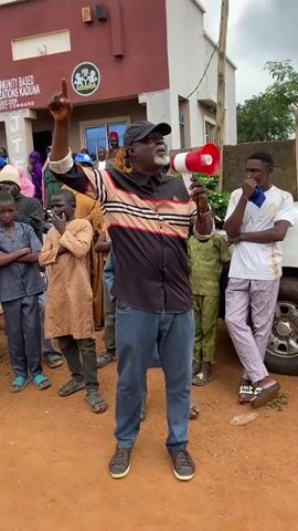 This media file depicts an outdoor gathering in Kawo, Nigeria. The central figure is a man wearing a black baseball cap, a patterned shirt with stripes of beige, black, and red, and blue jeans. He is holding a red and white megaphone and appears to be addressing a crowd. His right arm is raised, and his hand is open, suggesting he is speaking or making a point.

Surrounding the speaker are numerous individuals, including men, women, and children. Some are dressed in traditional attire, while others wear more casual clothing. Many of the children have their arms crossed, and some of the adults are looking towards the speaker. The overall atmosphere seems to be one of a public address or a community meeting.

Behind the gathering, a building with "COMMUNITY BASED ORGANIZATIONS KADUNA" and "NDC COMMAND" visible on its facade is present. The building has a red upper section and a light-colored lower section with windows and a door. There is a seal or logo on the building's front. A white vehicle is partially visible on the right side of the frame.

The ground is covered in reddish-brown soil, indicative of an unpaved area. The weather appears to be overcast or cloudy, and the lighting suggests it could be daytime, possibly late morning or afternoon, with no harsh shadows. The scene conveys a sense of community engagement and public discourse.