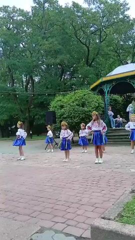 A group of young girls in traditional Ukrainian clothing are performing a dance in an outdoor park. The girls are wearing embroidered blouses, blue skirts, and floral headbands. They are arranged in a semi-circle facing a gazebo in the background. A speaker and a microphone are set up on a stand to the left of the girls. The ground is paved with cobblestones. There are trees and foliage in the background, and the sky is overcast. The girls are moving in unison, performing various dance steps and arm movements. Some of the girls are holding small ribbons in their hands. The performance appears to be for an audience, as there are people seated in the gazebo.