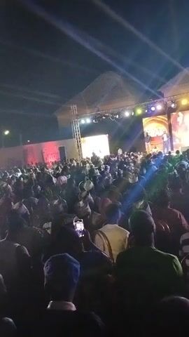 An audience has gathered at night for a performance or presentation, facing a stage that features two large video screens showing people on the stage. The audience members, mostly seen from the back, are diverse, with some wearing traditional hats or head coverings. Many are holding up their phones, presumably to record or take photos of the event. The stage is lit with a variety of lights, and there is a metal truss structure visible. The event appears to be taking place outdoors, under a roof or awning that is illuminated by the stage lights.