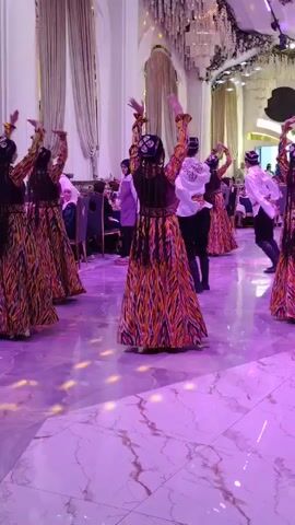 A group of women in traditional patterned dresses and head coverings are dancing in a ballroom. The women are performing synchronized movements with their arms raised. In the background, men and women are seated at tables, and some people are standing. The ballroom has large curtains, ornate chandeliers, and a marble floor that reflects the purple and pink stage lighting.