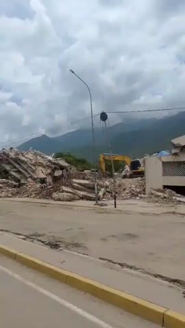 The video shows a pile of rubble from a destroyed building. A yellow excavator is visible behind the rubble, suggesting demolition or cleanup efforts. The scene is outdoors with a street in the foreground and mountains in the background under a cloudy sky. There are no people clearly visible in the frame.