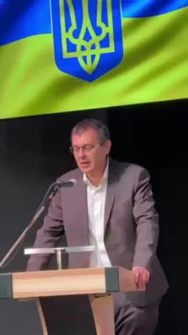 A man in a gray suit and glasses stands behind a light wood podium with a microphone. He is speaking. Behind him is a black wall with a yellow and blue Ukrainian flag hanging at the top.
