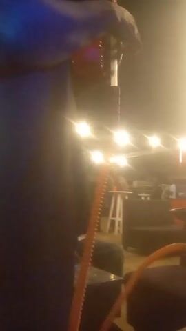 The video depicts a hookah lounge at night. People are sitting on couches and chairs, smoking hookah.  String lights illuminate the area. A person in a white sweater walks past a hookah on a table. Red plastic cups and bottles are visible on the table. The camera moves a bit during the filming.  Outside, the glow of illuminated signs is seen.