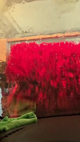 The view from inside a car going through a carwash is captured. The foreground features the car's dark dashboard with a green microfiber cloth resting on it. The camera is focused on the large, red brushes of the carwash as they move against the car's windshield, and large droplets of water obscure the view. The lighting inside the carwash cycles from red to pink to purple. Beyond the brushes, the opaque, slightly visible ceiling of the carwash can be seen.