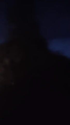 The video begins with a dark, cloudy sky. It then transitions to a view of a bright light, possibly a streetlamp, shining downwards and creating lens flares. In the foreground, a portion of a white vehicle, likely a car, is visible on the right side of the frame, with its red taillight illuminated. The overall scene suggests the video was recorded at night, possibly from inside a moving vehicle, given the motion blur and the presence of the car's interior elements.