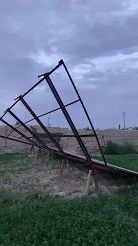 The video captures an outdoor scene in a rural or semi-rural area under an overcast, cloudy sky, suggesting either dusk or dawn. The primary subjects are a series of metal frames and partially installed solar panels.

The video begins with a close-up of a large, tilted metal frame structure, appearing to be made of brown or rusty-colored beams, resting in a field of green plants. This initial frame is skeletal. In the background, there are mounds of earth or debris.

As the camera pans to the right, more of the green field becomes visible. The pan reveals several rows of solar panels mounted on similar metal frames. The panels are dark, rectangular, and appear to be in various stages of installation or repair, some resting on makeshift wooden supports on the ground. They are laid out horizontally across the field.

In the mid-ground and background, there are scattered tall, slender trees (possibly cypress trees) and some distant, low-rise buildings or structures. The ground beneath the solar arrays is predominantly covered with short, green vegetation, mixed with patches of exposed soil or dirt. No people are visible, and there is no discernible text.

The scene depicts what appears to be a solar energy installation project, either under construction, incomplete, or damaged. The location is Qila Saifullah, Pakistan.
