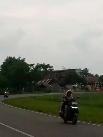 The video shows a rural landscape under a cloudy sky. A paved road curves through the scene, bordered by a grassy area and a fence. On the right side of the road, there are some buildings, trees, and what appears to be a field with some flags or decorations. On the left side of the road, there are more trees and some houses. A motorcycle with two people on it is seen riding on the road in the first few seconds. In the distance, a mosque with a dome and a minaret can be seen. There are also some parked motorcycles near the buildings on the right. The overall impression is of a quiet, rural setting.