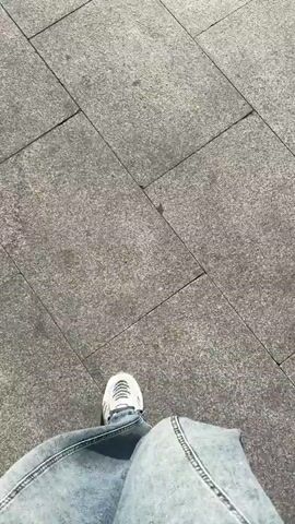 The video begins with a close-up shot of a person's feet walking on a tiled sidewalk. The camera then pans up to show a street scene with cars and motorcycles at an intersection. A pedestrian crossing is visible, as are traffic lights and street signs. The sun is shining, suggesting it is daytime. The architecture in the background appears to be urban. The video then cuts back to the street scene, showing more cars and motorcycles moving through the intersection. The footage is shaky, indicating it was likely filmed by someone walking or riding a bike.