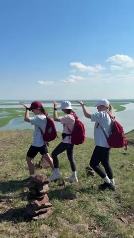 The video shows three young women walking in single file up a grassy hill on a sunny day. They are all wearing casual clothes and backpacks. The woman in the front is wearing a red baseball cap, a white t-shirt, black shorts, and sneakers. The woman in the middle is wearing a white baseball cap, a pink t-shirt, black leggings, and white sneakers. The woman in the back is wearing a white baseball cap, a light blue t-shirt, black leggings, and black sneakers. They are all carrying red backpacks. The woman in the middle is touching the woman in the back's backpack. They are walking away from the camera, towards a body of water with green vegetation. There are stacks of rocks on the ground in the foreground.