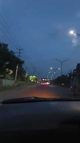 The video shows a view from inside a car as it drives down a road at dusk. The sky is a deep blue, and streetlights are illuminated. There are utility poles with wires running along the side of the road. Trees and some vegetation are visible. The road has lane markings, and there are signs and traffic signals. Other vehicles, including a bus or large truck, are on the road ahead. The dashboard of the car is visible in the foreground. The overall atmosphere is that of a nighttime or twilight drive on a suburban or rural road.