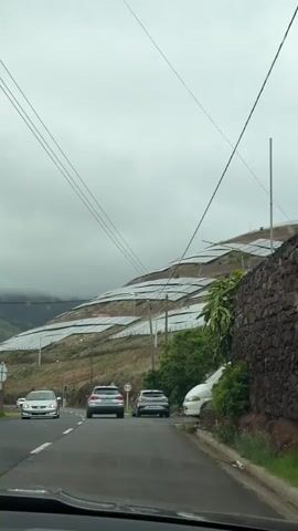 The video shows a view from inside a car, looking out at a road and a hillside. The road has three cars on it, and the hillside is covered in what appear to be rows of greenhouses or some kind of structure covered in translucent material. There are telephone poles running along the side of the road, and a sign with the number 60 on it is visible. The sky is overcast, and the overall impression is of a rural, somewhat gloomy scene.