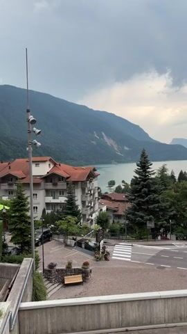 The video captures a scenic view of a town nestled beside a large lake, surrounded by lush green mountains. The sky is overcast with dramatic clouds. In the foreground, a street corner with a pedestrian crossing is visible. A few cars are present, with one black car driving on the road. Buildings with terracotta roofs are scattered throughout the town. Tall evergreen trees dot the landscape, adding to the natural beauty. A concrete barrier runs along the bottom of the frame, suggesting the viewpoint is from an elevated position. The overall impression is one of a peaceful, picturesque mountain town.