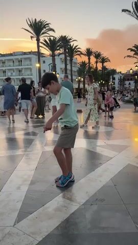 A young boy is dancing in the middle of a large paved area. He is wearing a light green t-shirt, beige shorts, and blue sandals. He is in focus and centered in the frame. He is bending at the waist and his arms are out to his sides. The paved area has a pattern of light and dark stone tiles. In the background, there are several palm trees, buildings with balconies, and other people walking around. The sky is a warm orange and pink color, indicating it is sunset. The lighting suggests it is dusk.