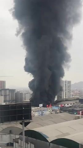 A plume of thick black smoke billows into the sky from a building in a city. The smoke rises high above surrounding buildings, obscuring much of the sky. Flames are visible at the base of the smoke plume, indicating a fire. The buildings appear to be a mix of residential and commercial structures. The overall scene suggests a significant urban fire.