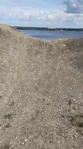 The video captures a first-person perspective of walking towards the edge of a cliff or hill overlooking a large body of water. The foreground consists of rough, greyish-brown ground covered with small rocks and gravel, with sparse green vegetation. As the camera moves forward, the expansive water body comes into full view. The water is dark blue, with gentle ripples reflecting the daylight. In the distance, across the water, a green tree line is visible, along with several buildings, suggesting a city or town on the far bank. The sky is partly cloudy with patches of blue. The overall scene depicts a natural landscape, likely a viewpoint overlooking a wide river or lake, consistent with the provided location context of "Пятый километр, Russia". No people or visible text are present in the video.