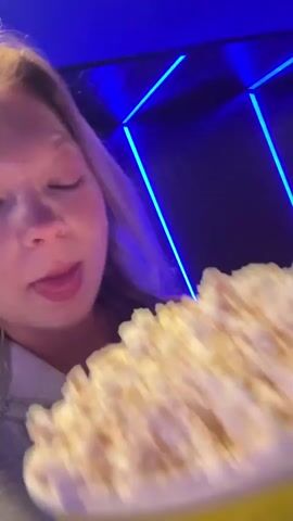 A woman is eating popcorn in a movie theater. She is holding a large bucket of popcorn and shoving handfuls into her mouth. The theater is dimly lit, with neon lights in the background. The woman is wearing a hooded sweatshirt and a necklace. She makes a kissy face and glances around the theater.