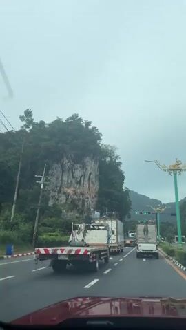 The video is a first-person view from inside a vehicle, looking forward. The vehicle is on a road with multiple lanes, with other vehicles including trucks and cars on the road. The road appears to be a highway, with a large, rocky mountain covered in trees on the left side of the frame. The sky is overcast and appears to be a dull, grayish color. There are several ornate lampposts with decorative gold elements lining the right side of the road. In the distance, there are more mountains and lush green vegetation. The traffic appears to be moving, though it is not fast-paced. There are traffic lights visible, showing green. The overall impression is a scenic drive through a mountainous area with some traffic.