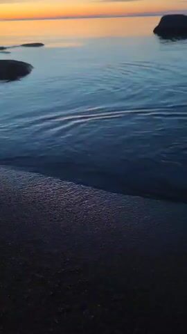 A close-up, low-angle shot shows a small wave washing onto a dark, sandy shore during sunset. The water is a deep blue, reflecting the soft oranges and yellows of the sky. Several dark rocks are visible in the distance, partially submerged in the calm water. The camera is focused on the gentle movement of the water as it recedes from the sand.